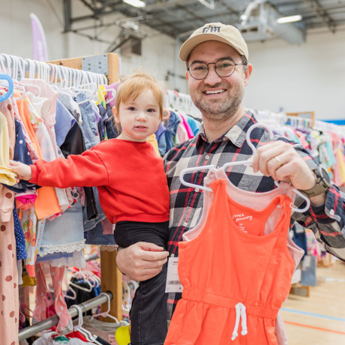 A mom carrying her baby in a front carrier holds a purple JBF bag and a hanger with denim overall shorts. She is standing next to a rack of baby clothing.